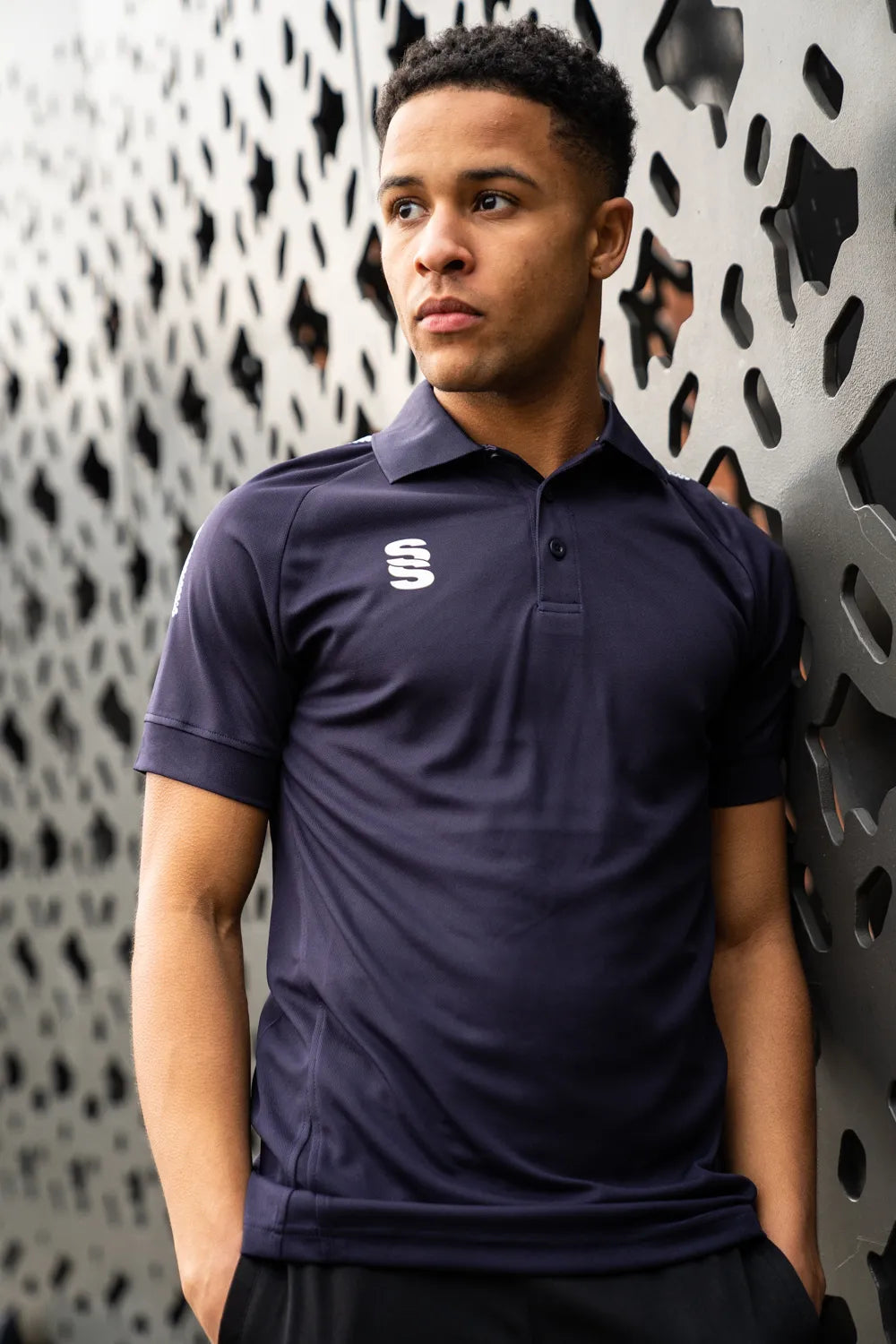 A person wearing a white stuart surridge sports polo shirt with a solid color design and a logo on the right chest area, standing in front of a patterned wall.