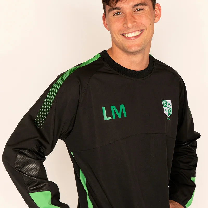A smiling man wearing a black long-sleeve athletic top with green accents, including green mesh panels on the shoulders, green 'LM' initials on the chest, and a green and white shield crest on the left chest