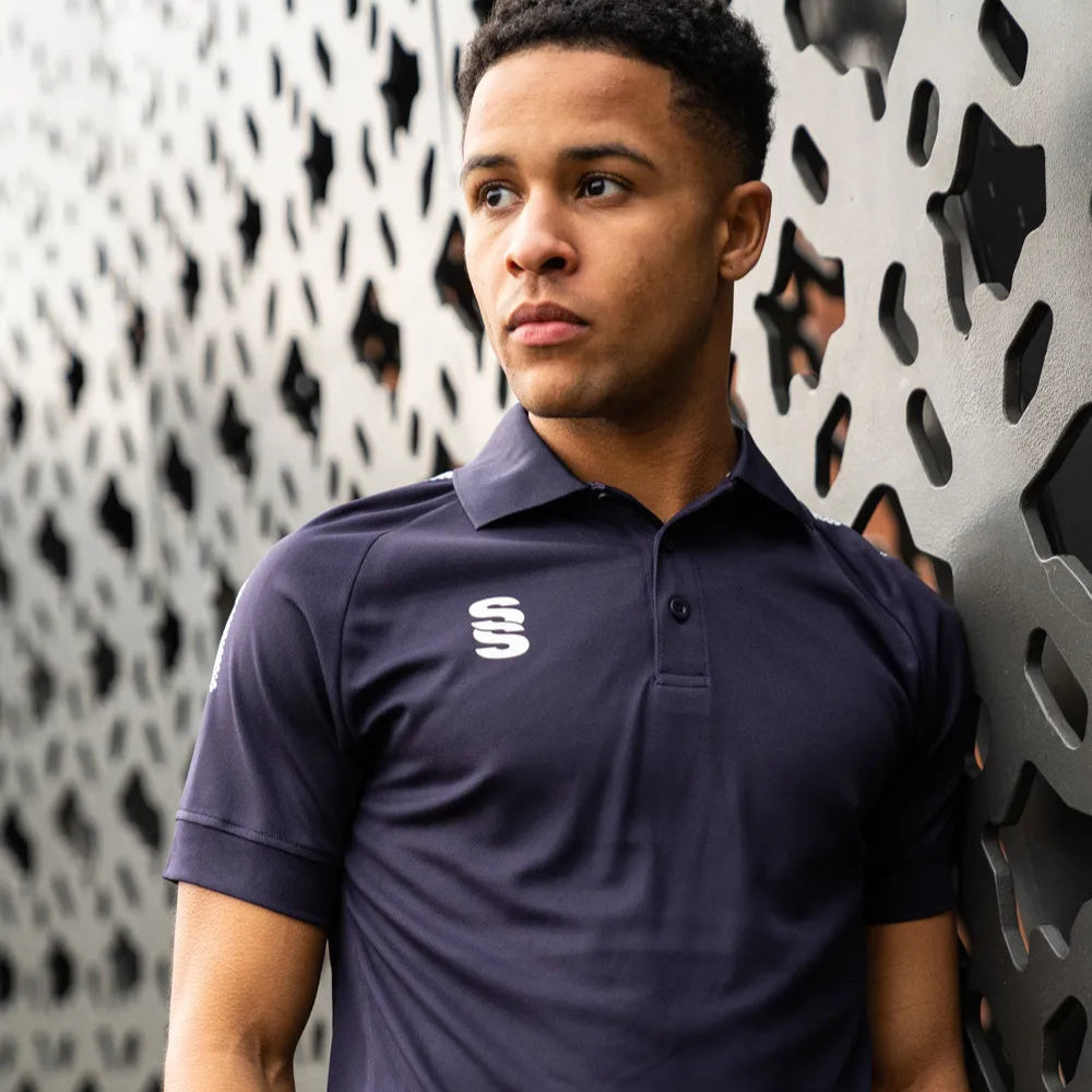 A person wearing a white stuart surridge sports polo shirt with a solid color design and a logo on the right chest area, standing in front of a patterned wall.
